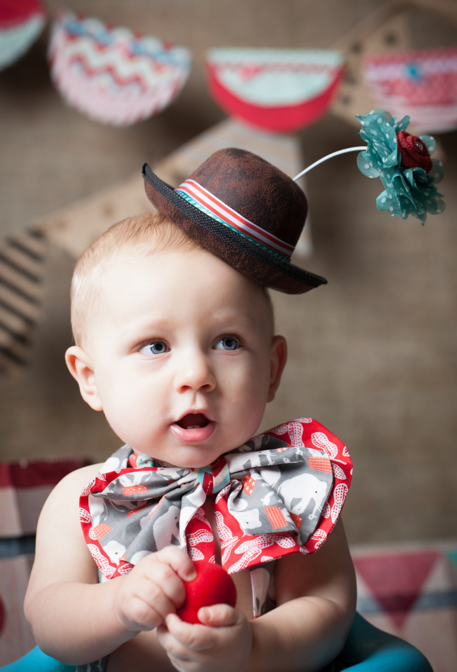 Little boy in brown hat with a big silly flower and an oversized patterned bow with a circus backdrop for a birthday party event photography