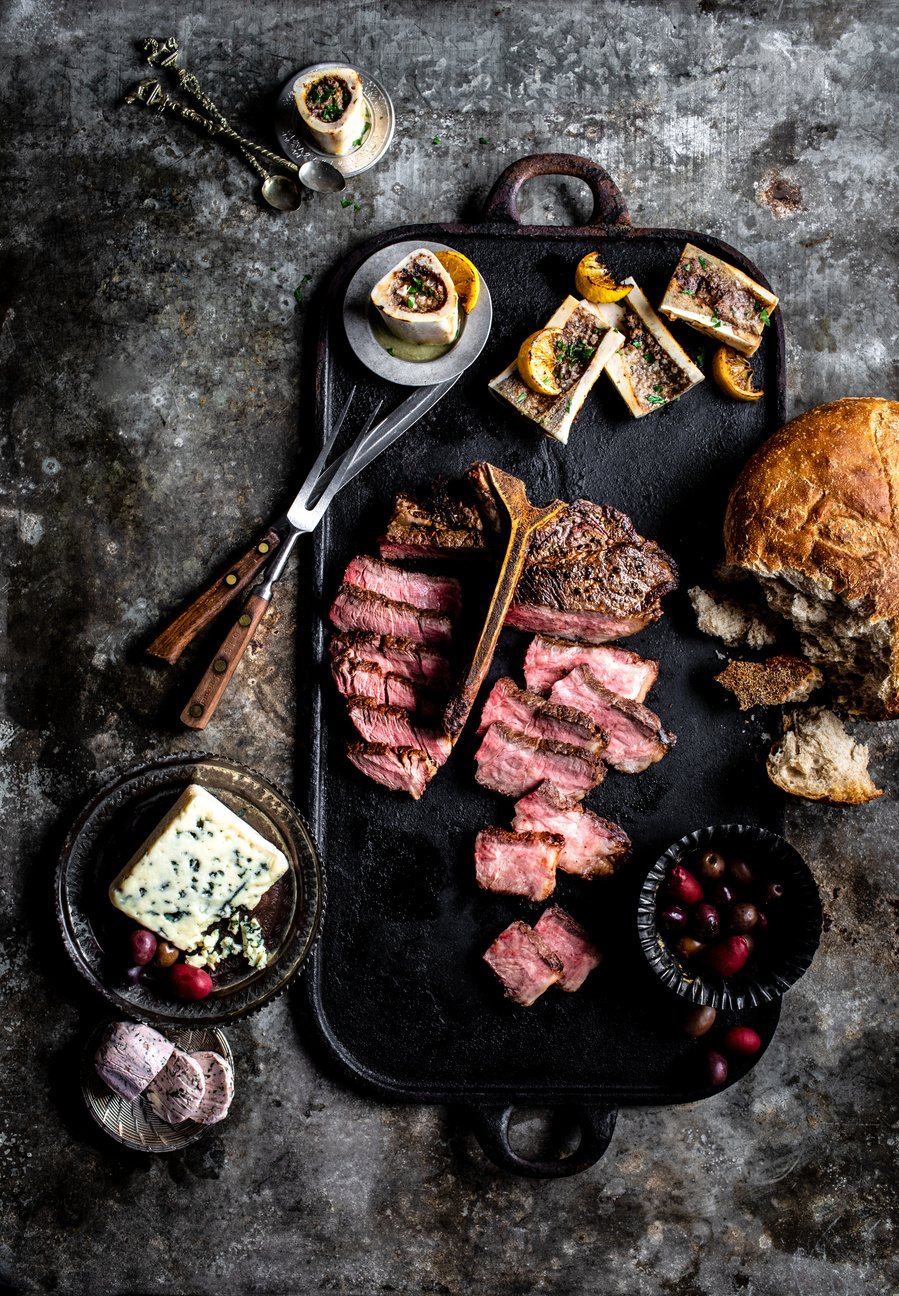 Overhead meat photography spread of T-bone steak sliced on a tray with bread, cranberries, and blue cheese surrounding. 