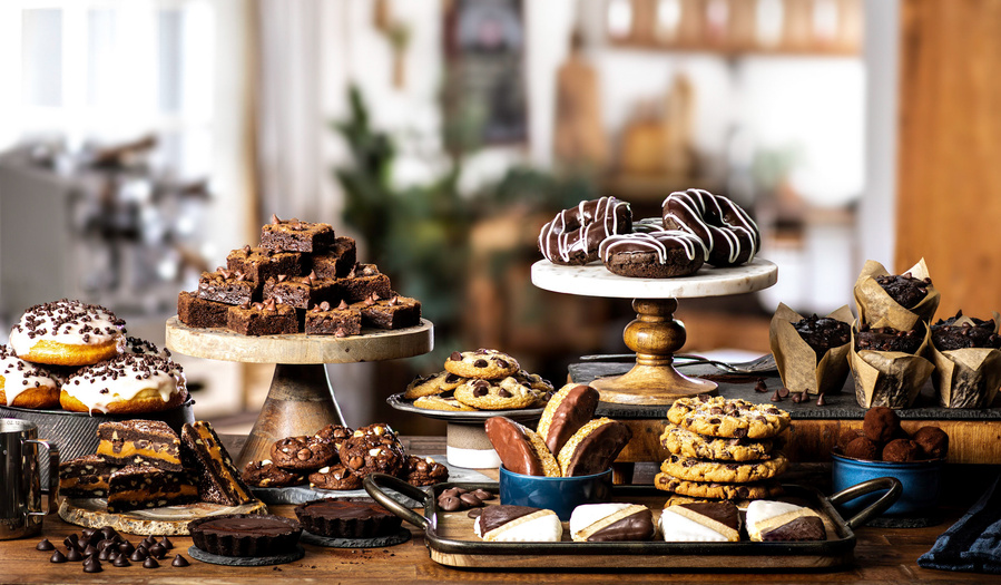 Elaborate chocolate treat table display with doughnuts, cookies, and muffins stacked in multi-level Ghirardelli chocolate photography spread. 