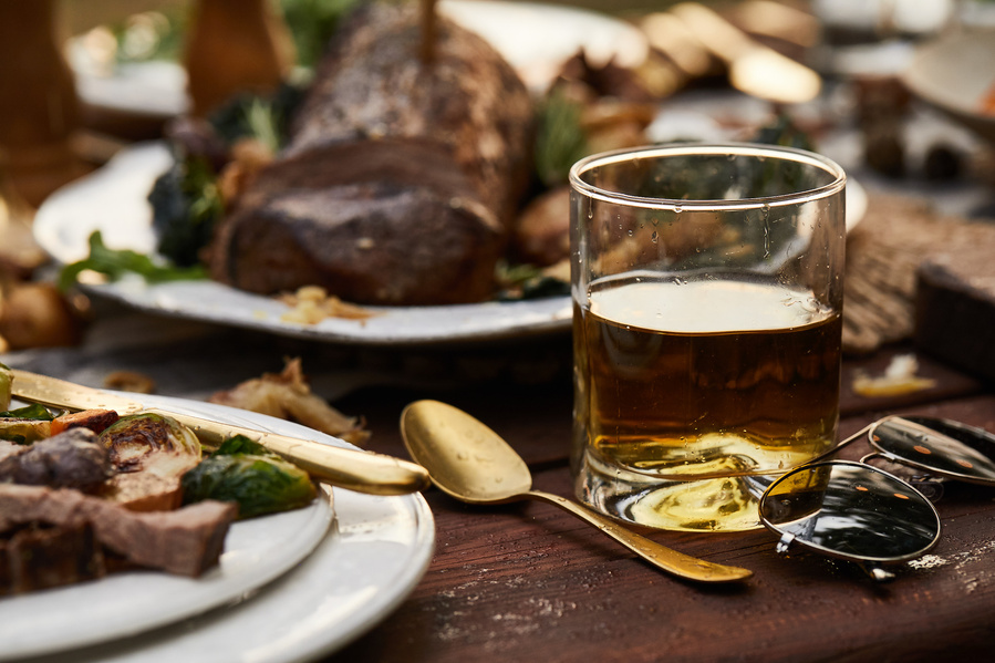 Tight shot of glam outdoor dinner party picnic table with roasted meat, plated dinner and glass of amber alcohol with gold spoon. Lifestyle food photography for The Buckle