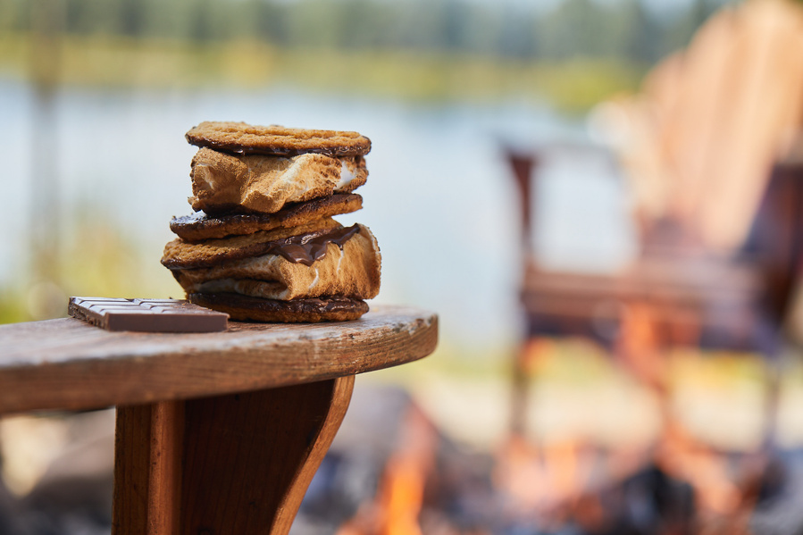 Double decker smore sitting on the wooden arm of a chair around a firepit on the lake shore. Lifestyle food photography for the Buckle