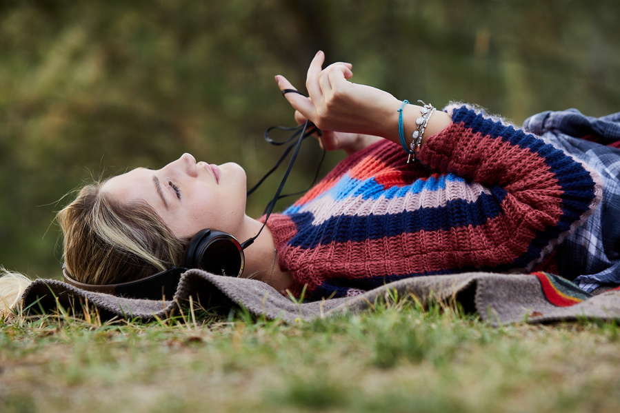 Young woman laying on a blanket in a striped sweater listening to music on big headphones and playing with the cord. Fashion lifestyle photography for The Buckle