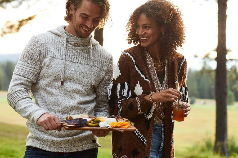 Two fashion models in rustic fall clothes holding charcuterie board and cocktail as they sit down to outdoor dinner party for lifestyle food photography