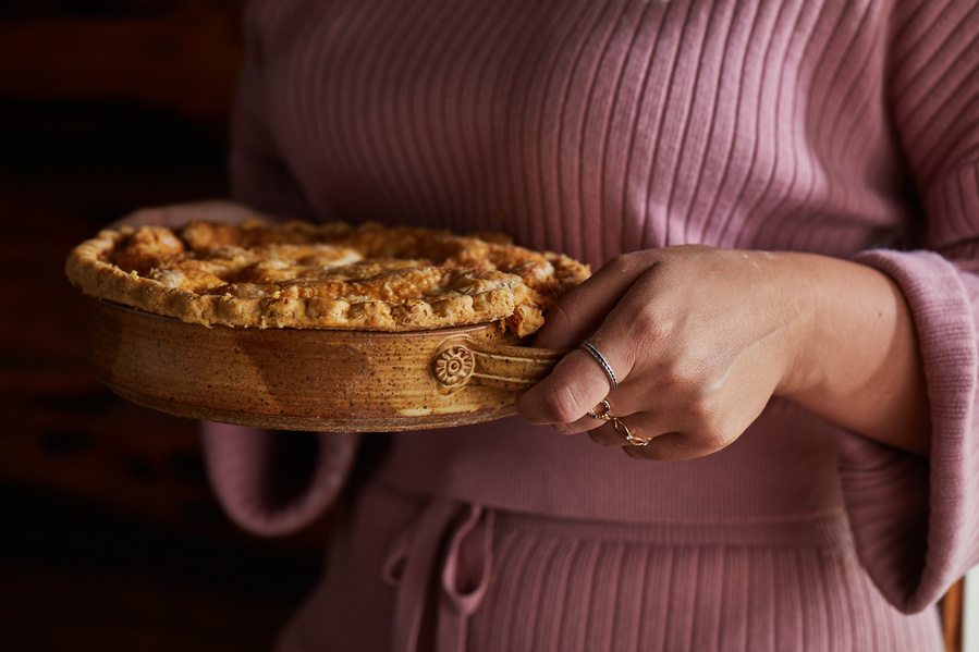 Woman in pink sweater and shorts set holding a baked pie in a dish. Lifestyle food photography for The Buckle