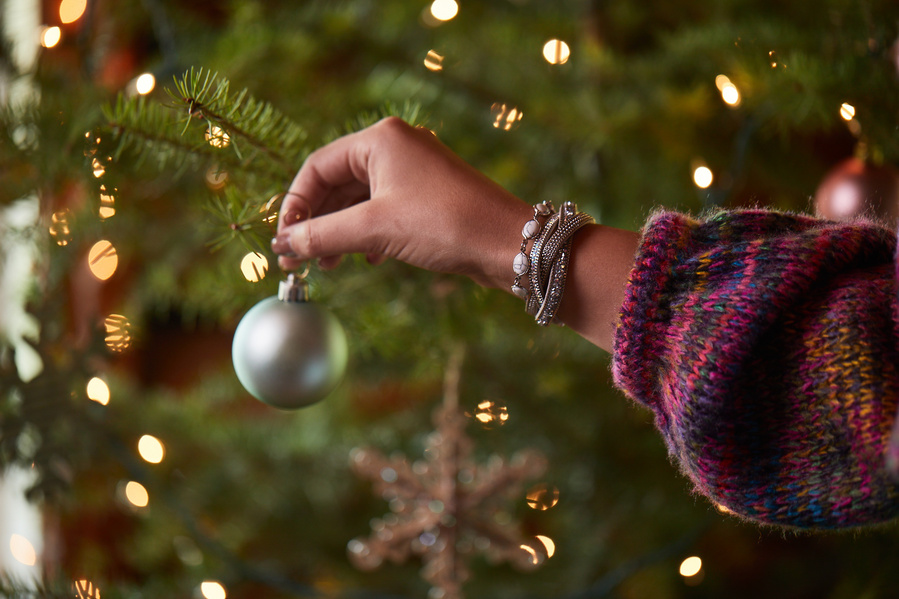 Woman's hand in red and blue sweater and bracelets hanging a silver ornament on a lit Christmas tree. Fashion lifestyle photography for The Buckle