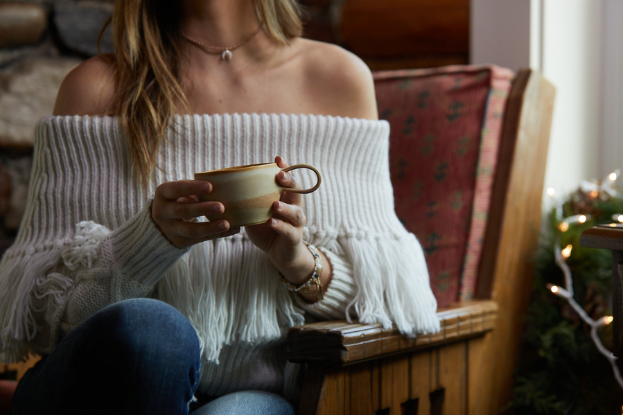 Female fashion model in off the shoulder gray sweater holding a mug in a cabin chair near a window. Fashion lifestyle food photography for The Buckle