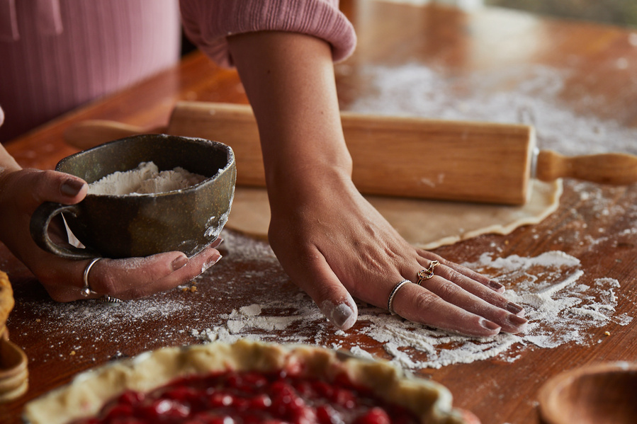 Woman's hand spreading flour on a table top with pie crust and rolling pin in background and cherry pie in foreground. Fashion lifestyle food photography for The Buckle. 