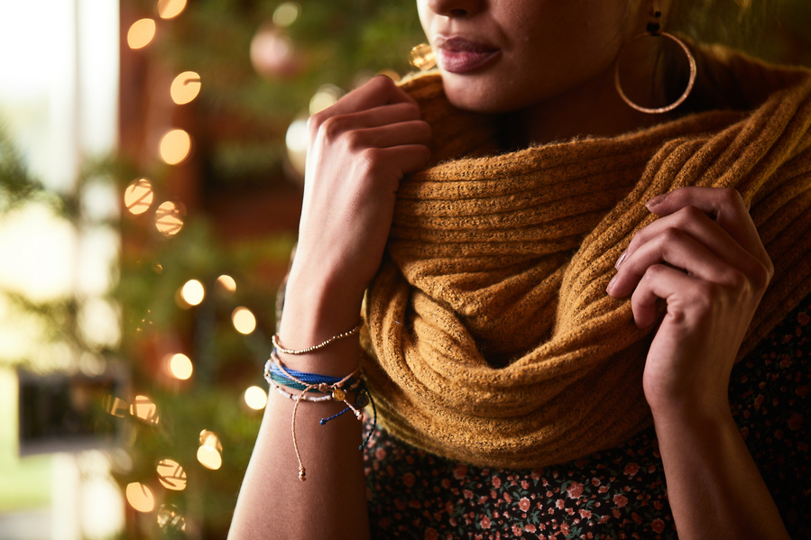 Model poses in front of lit Christmas tree with hands on large yellow cowl scarf. Also wearing bracelets and hoop earrings. Fashion lifestyle photography for The Buckle