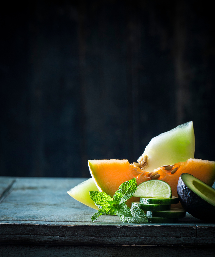 Orange and green melon slices standing on the rind with other produce in front. Sitting on a blue wood surface with a dark background and beautiful light. Used as cover for photographer profile in Click Magazine