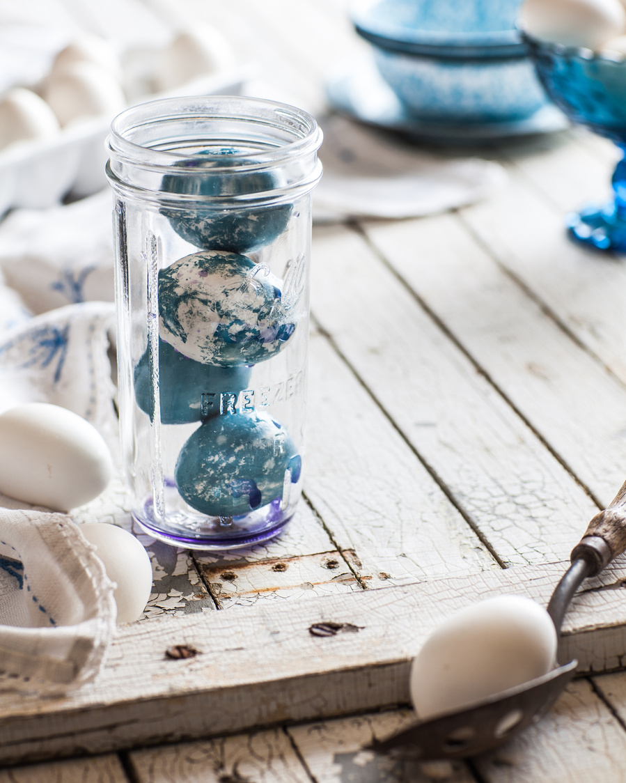 Four blue and white dyed easter eggs stacked in a narrow mason jar on a white wood surface with accessories around for dyed egg photography
