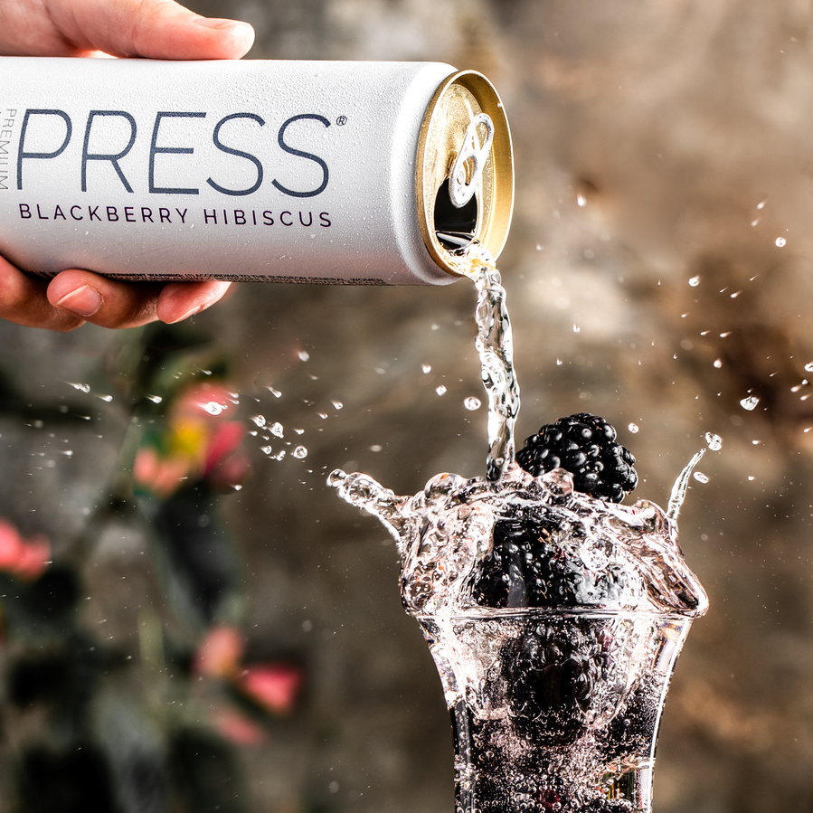 Pour and splash drink photography of can of Press Seltzer pouring into glass with blackberries. Hibiscus bush in the background. 