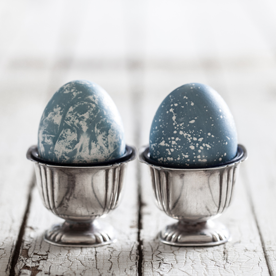 Two blue and white speckled eggs in shiny silver egg cups on white wood surface. Blue cabbage dyed eggs 