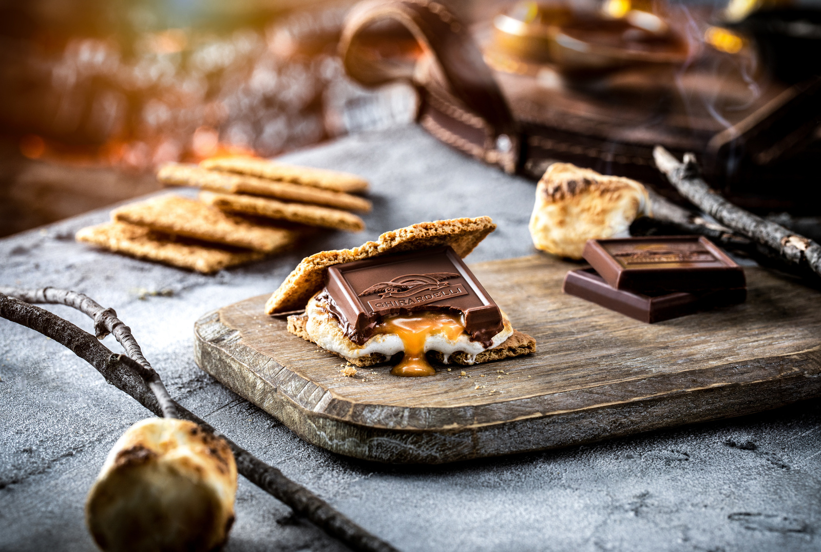 Chocolate, marshmallows, and graham crackers on a picnic table near a fire. A s'more is in the middle of the frame with a Ghirardelli caramel chocolate dripping down. Chocolate photography ad.