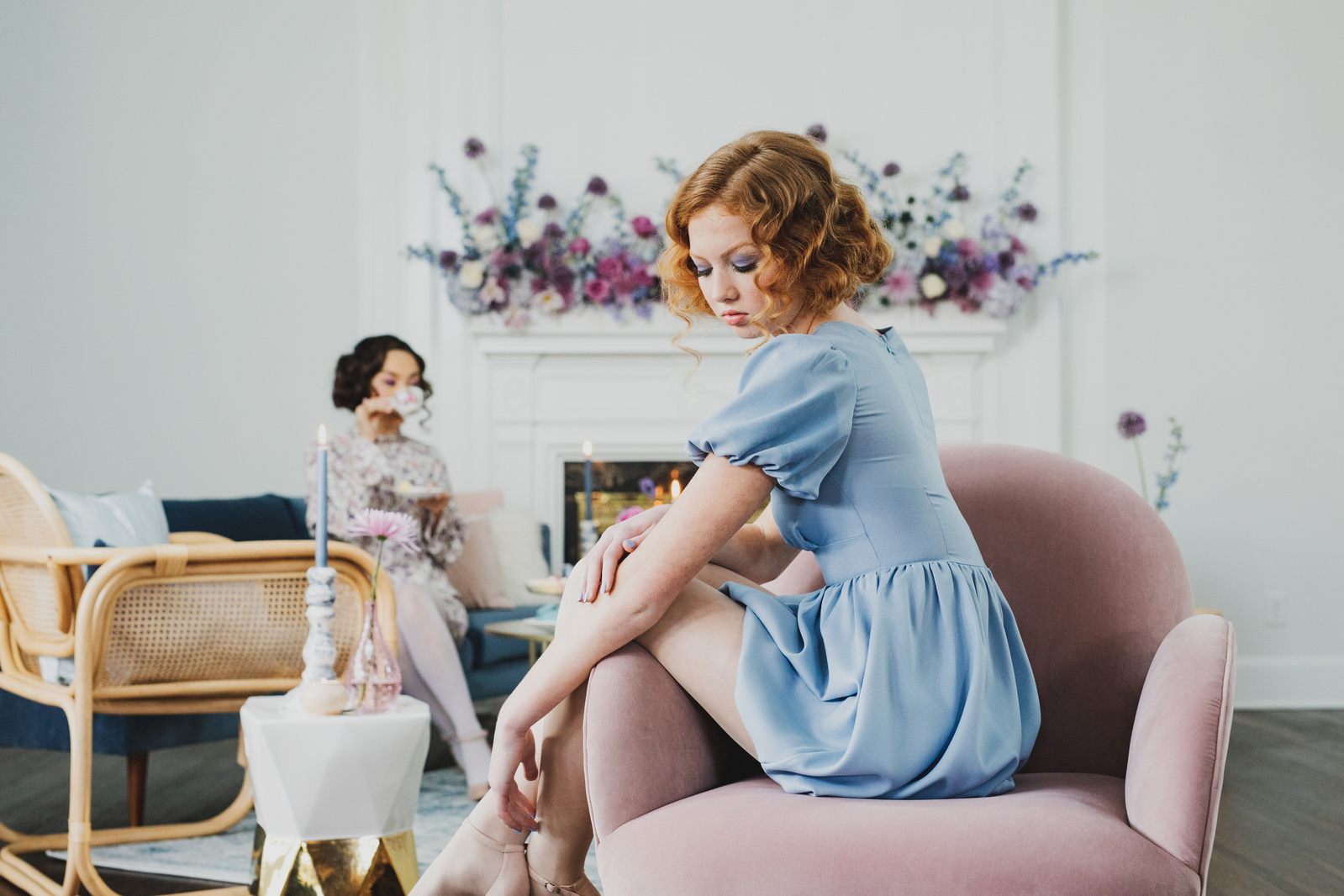 Two dainty models with bobbed hair sit in a white room with mauve accents and flowers. One sips tea in the background with a table for dessert food photography
