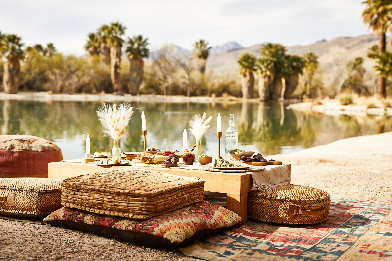 Glam picnic set up on a blanket with cushions on a sandy lakeshore with palm trees in the distance. Lifestyle food photography for The Buckle