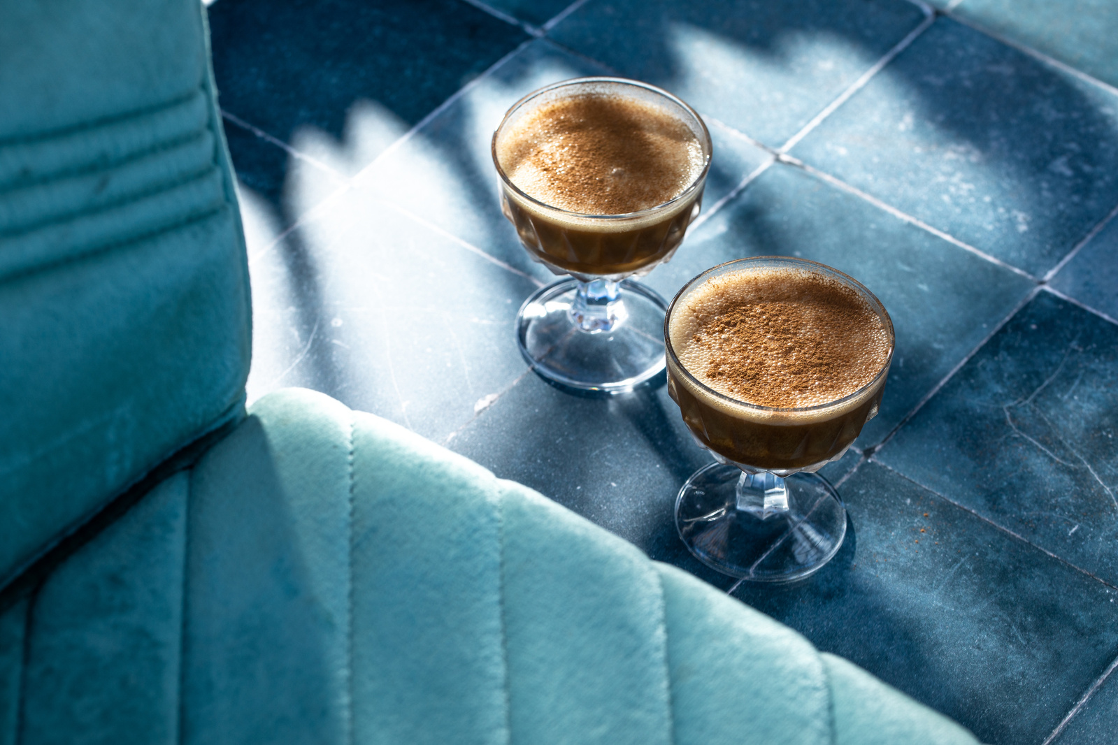 Restaurant photography with two espresso martinis on a teal tile surface next to a lighter teal seat with dramatic hard light and shadows