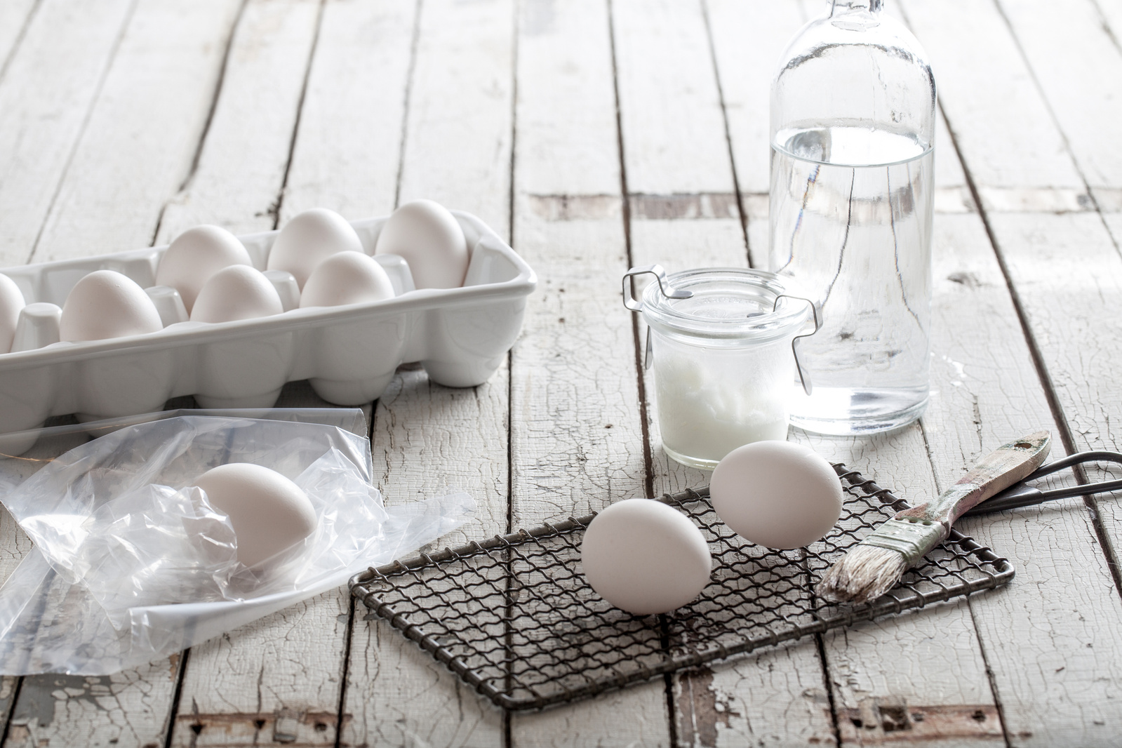 white eggs in a carton and on a grid with dying supplies on a white wood surface. setup image for diy cabbage dyed eggs photography