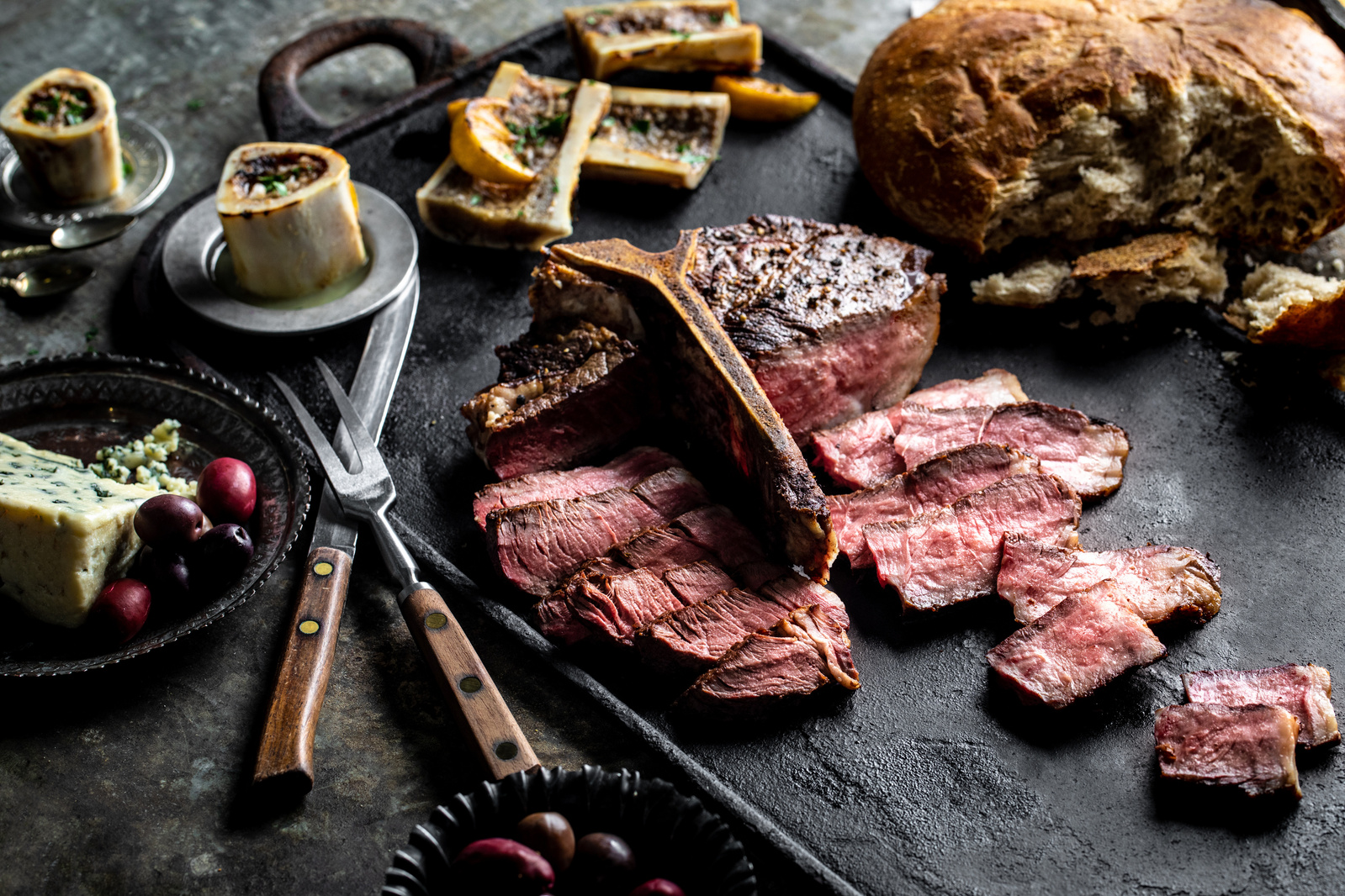 Food meat photography of a slices medium rare T-bone steak on slate with utensils, bread, and other props. 