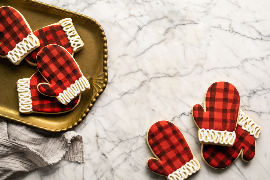 Four buffalo print mitten cookies on a bronze tray to the left with three more mitten cookies on the marble table to the right. Baking photography shot overhead with room left for editorial copy. 