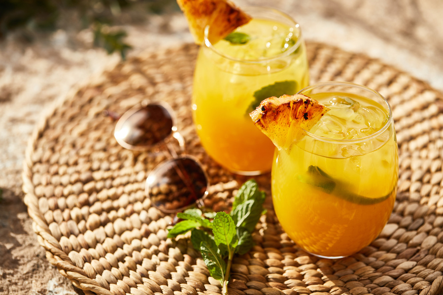 Two orange cocktails with pineapple garnish on a woven place mat with sunglasses in hard sunlight. Lifestyle food photography for The Buckle