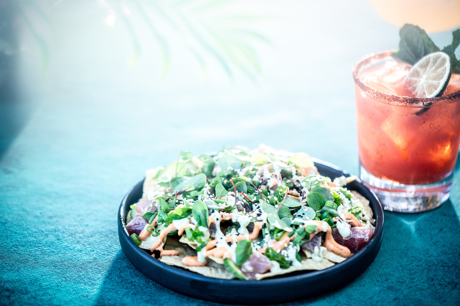 A black plate of poki nachos on a teal surface in bright hazy sunlight with a palm overhead. Bloody Mary sits to the right for restaurant photography. 