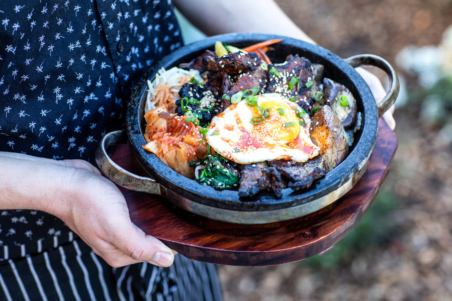 A cast iron dish of bibimbap on a wood trivet held with two hands by a chef as part of a restaurant food photography campaign