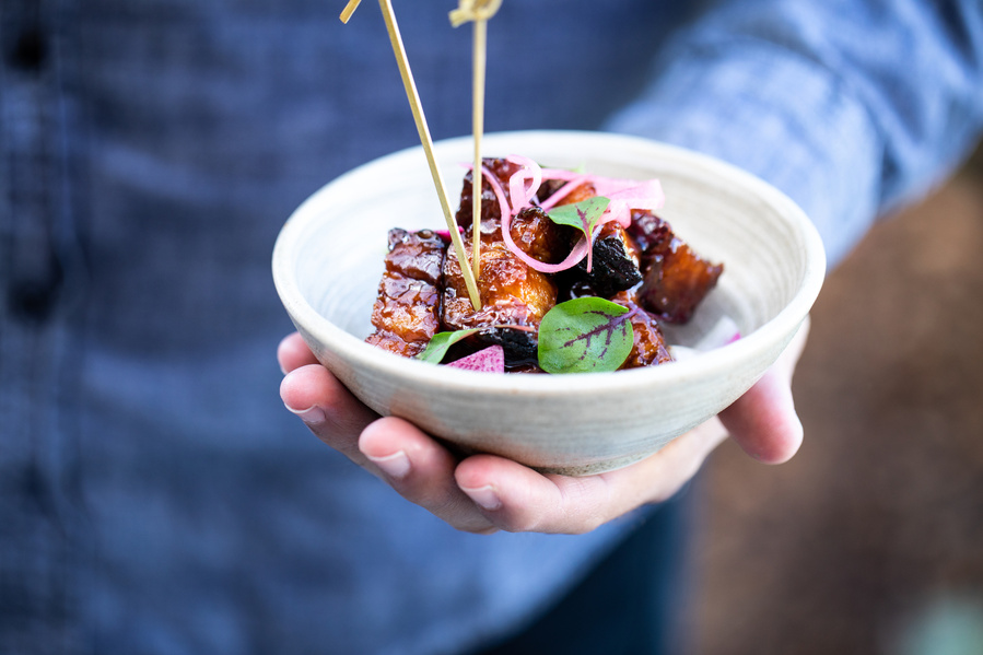 A white bowl held in the palm of a man's hand with caramelized pork belly appetizer with skewers for restaurant photogrpahy
