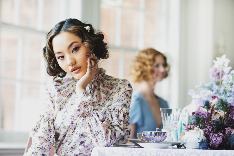 An editorial photography image capturing the whimsy and charm of a Bridgeton style tea party. One model in floral top with chin in hand looking at the camera and second model at the table behind. Feminine charm with vintage flare.