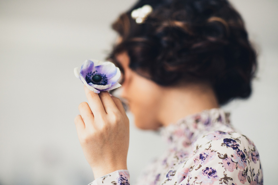 Editorial photography of model in floral high neck top with brown bobbed curls hiding her turned face with a purple flower. 