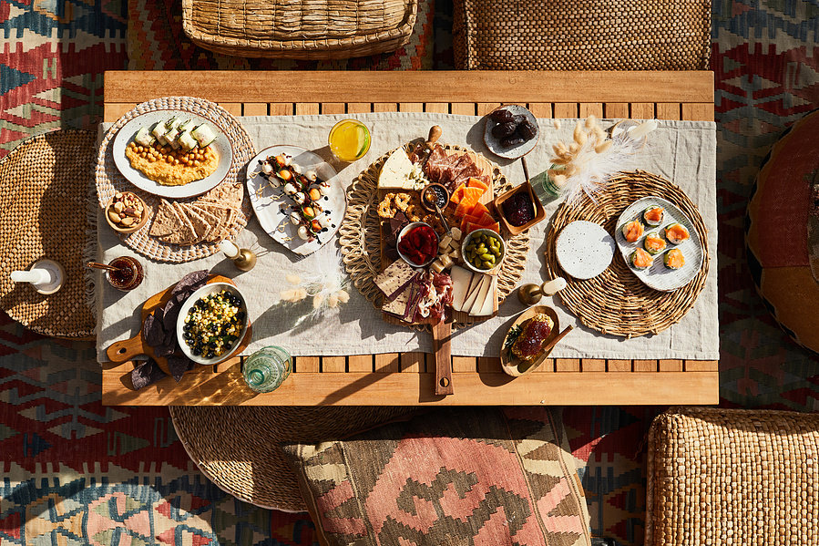 Overhead shot of short rectangle table with spread of snacks, charcuterie, and cocktails  on a blanket surrounded by cushions. Hard sunlight and long shadows in lifestyle food photography for The Buckle