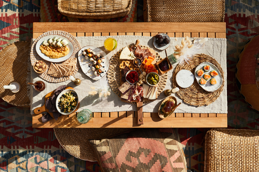 Overhead shot of short rectangle table with spread of snacks, charcuterie, and cocktails  on a blanket surrounded by cushions. Hard sunlight and long shadows in lifestyle food photography for The Buckle