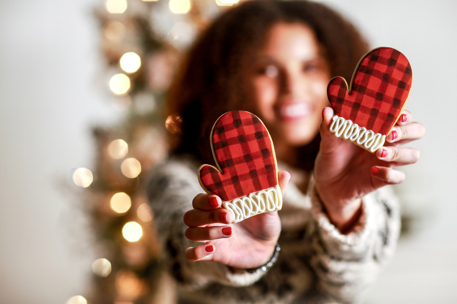 Model with curly brown hair holds two buffalo plaid mitten cookies out in front of her, framing her face with twinkle lights in the background. The cookies are in focus and she is not. 