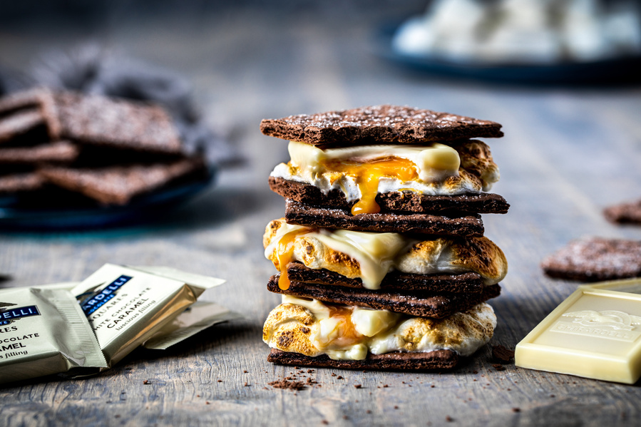 Triple layer s'more with chocolate graham crackers and white chocolate caramel Ghirardelli squares. Straight on chocolate photography with more wrapped chocolates on the table. 