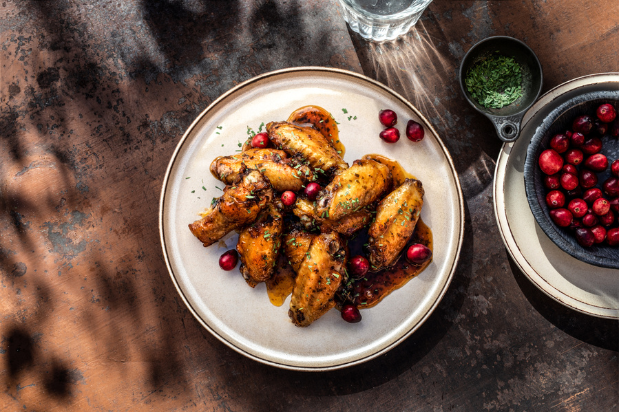 A creative food photography image featuring a stoneware plate of chicken wings with cranberry bbq sauce on a rustic surface. Dish of fresh cranberries  and parsley to the side and glass with sun shining through it. 