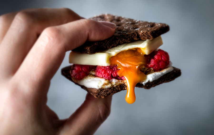 Close up of a hand holding a s'more with a bite out of it. S'more has chocolate graham crackers, white chocolate caramel Ghirardelli squares, and raspberries. Chocolate photographer caught the caramel dripping.