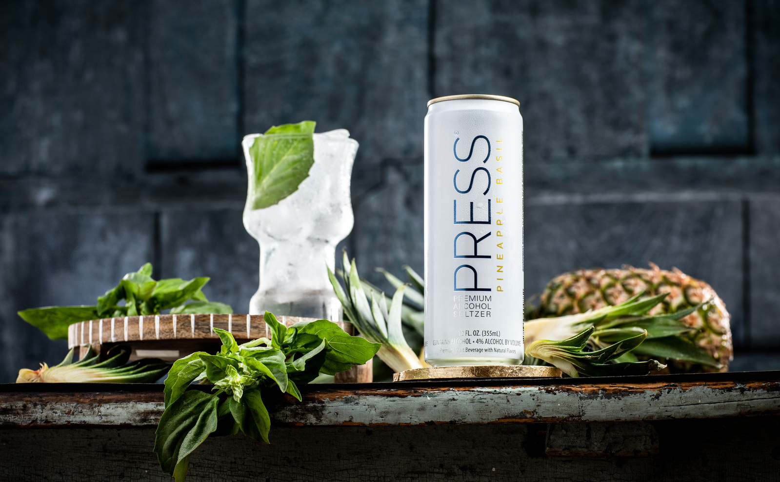drink photography of a glass of press seltzer next to the can with basil leaves and pineapples against a dark backdrop. 
