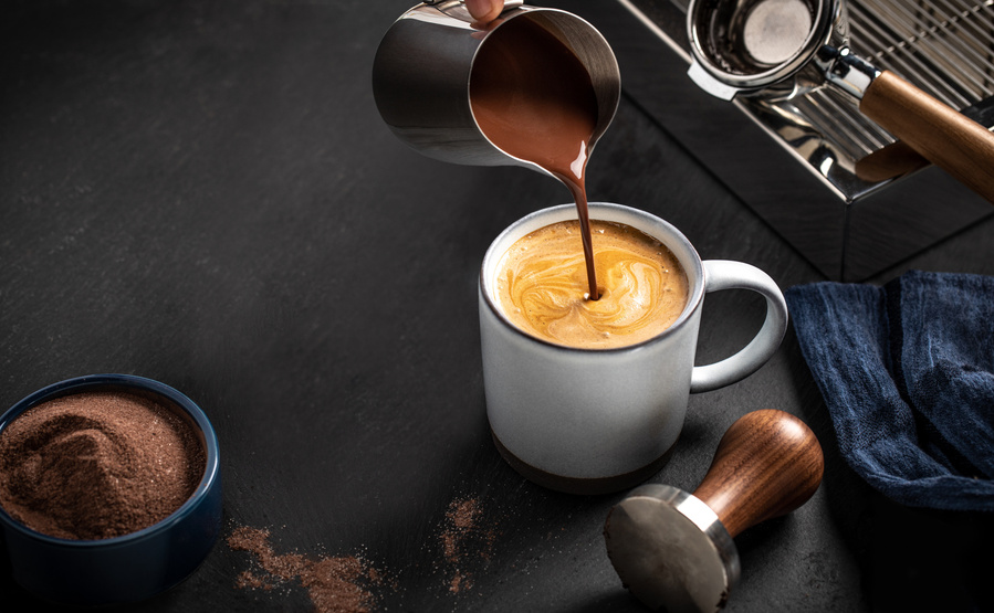 Slow pour of melted chocolate from a metal container into a latte in a large white mug. Looking into mug from above with moody chocolate photography lighting.