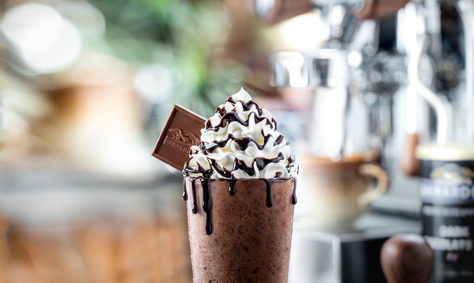 Frozen chocolate mocha with whipped cream, chocolate sauce, and Ghirardelli square in front of a shiny espresso machine.  Artistic chocolate photographer style. 