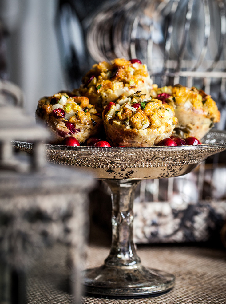 A grouping of cranberry muffins on a shiny silver cake stand surrounded by other shiny silver props. High contrast food photography.