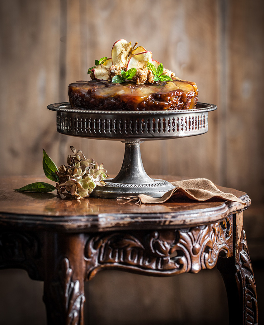 Apple cake on a tin cake stand on a ornate wooden table with apple blossoms and an apple topping. Cake photography straight on. 