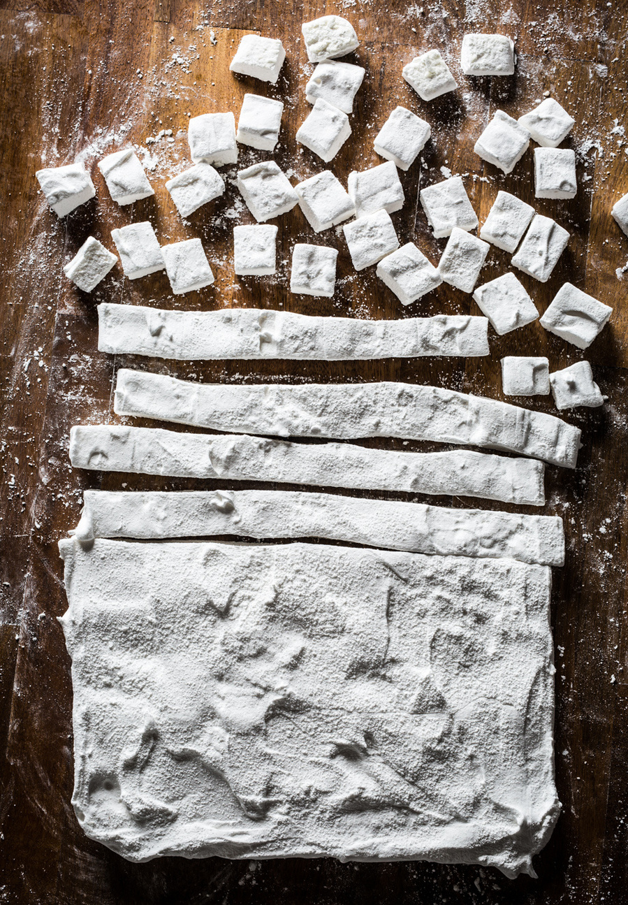 Overhead shot of pan of homemade marshmallows being cut into rows and then cubes on a wooden surface. Getting ready for drink photography of hot chocolate. 