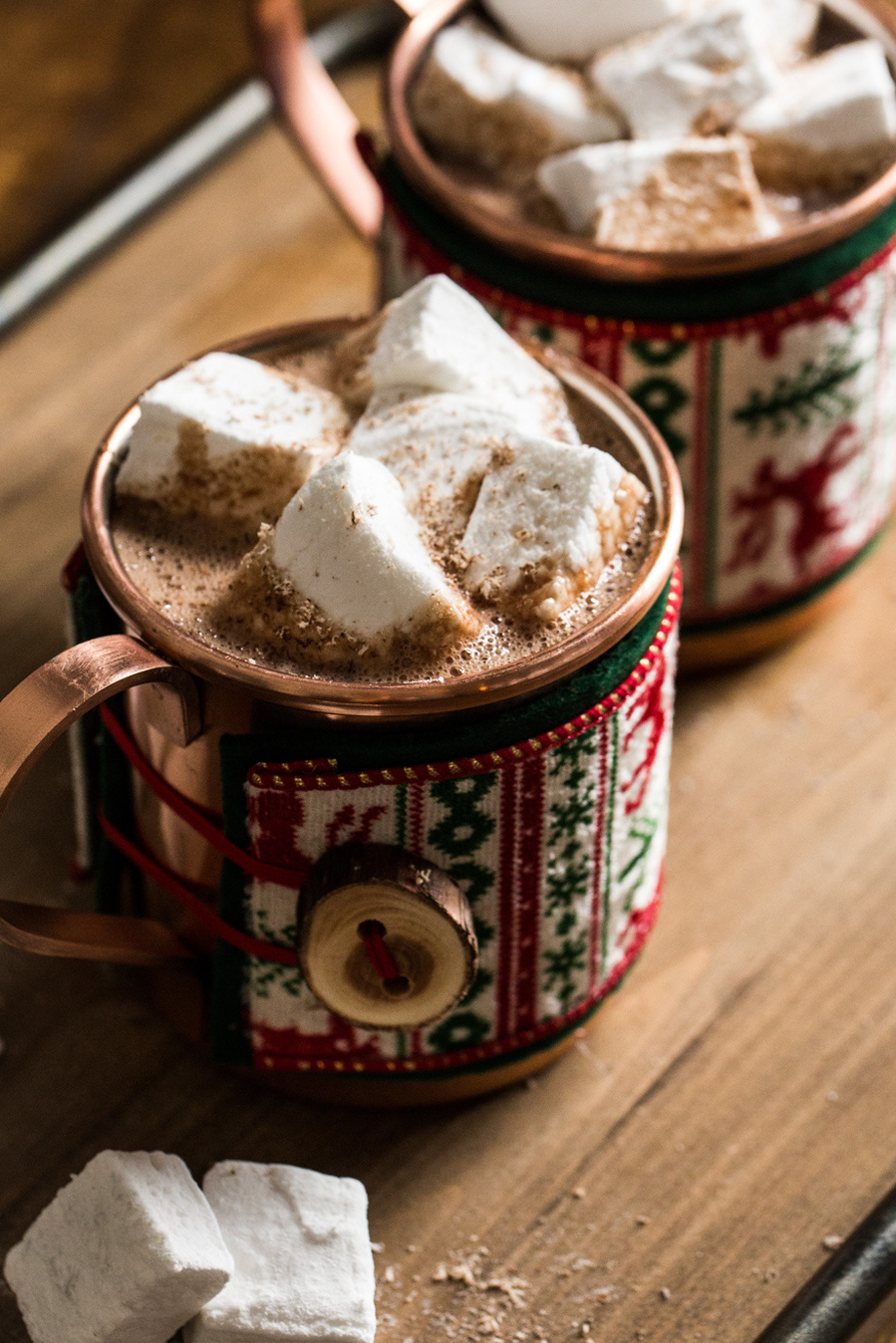 Two copper mugs of hot cholate topped with homemade marshmallow cubes. Mugs are wrapped in Christmas sweater cozies for drink photography 