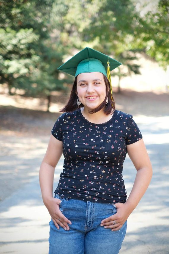 White teenage girl wearing black top, blue jeans, and green graduation cap, smiling with hands in pockets, with trees in the background