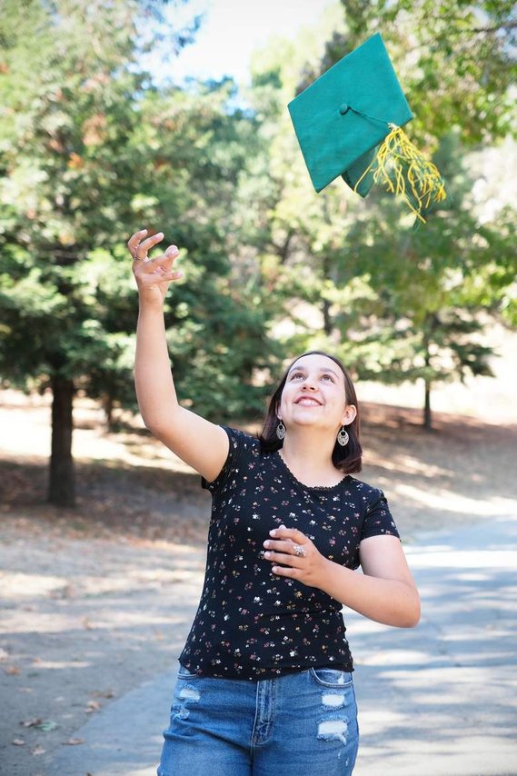 White teenage girl in black top and jeans, throwing green graduation cap up in the air and looking up at it, with trees in the background