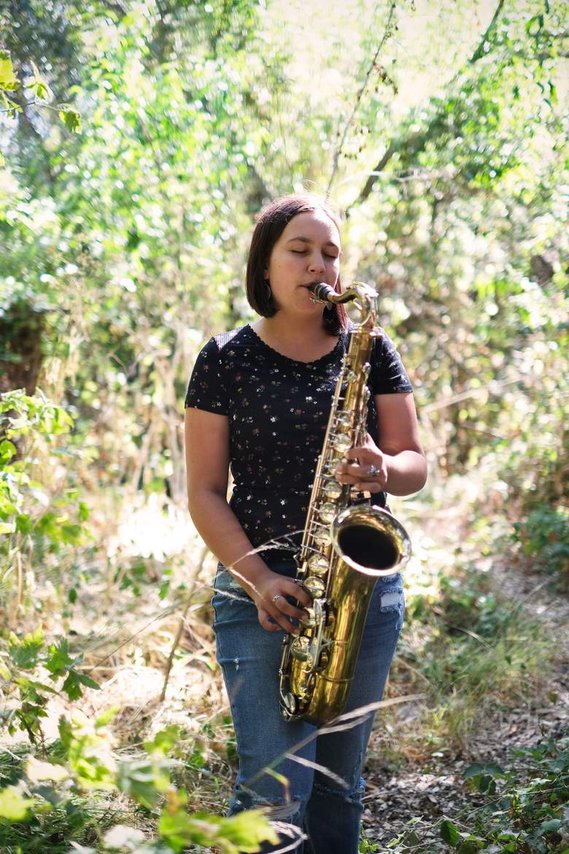 White young woman with short brown hair standing amidst trees playing saxophone with her eyes closed, wearing black top and blue jeans