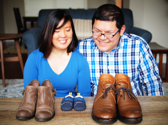 Asian couple sitting in living room, both looking down at coffee table with three pairs of shoes, one pair men's boots, one pair women's boots, and one pair baby shoes and smiling