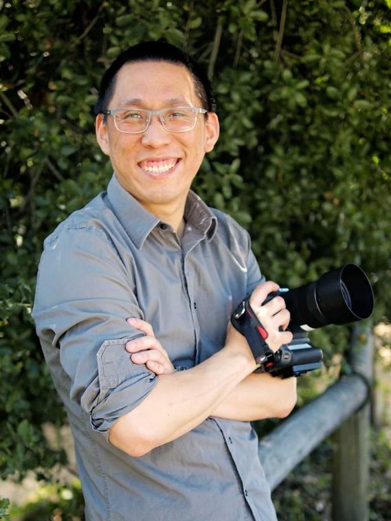 Asian man in gray collared shirt with glasses, holding camera in his hand, leaning against a wooden rail and smiling