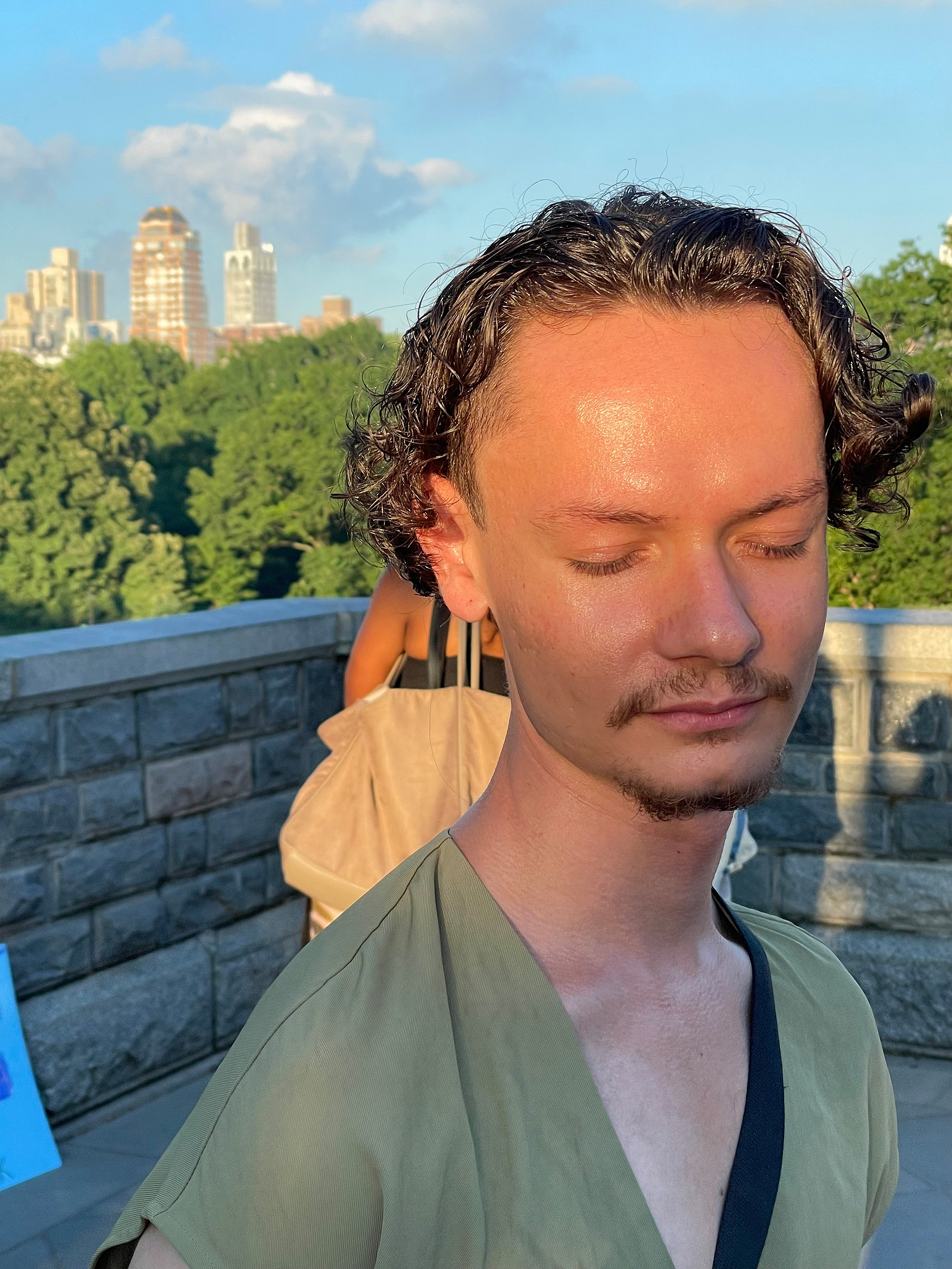 Anthony in the summer for 2022 at Central Park during CBU's Shakespeare in the Park. The sun is shining on Anthony's face. Anthony has his eyes closed. Some buildings of NYC are in the background behind some trees. Anthony is on the Belvedere Castle