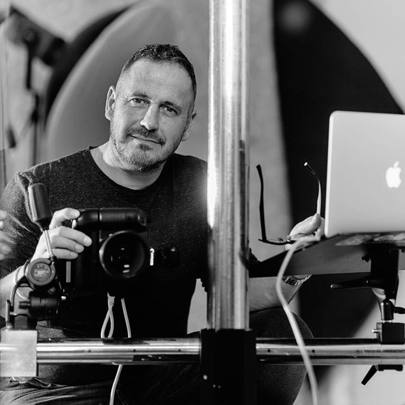 Black and white portrait of a commercial photographer, Matt Keal, holding his camera and Apple Macbook in his Norwich photography studio.