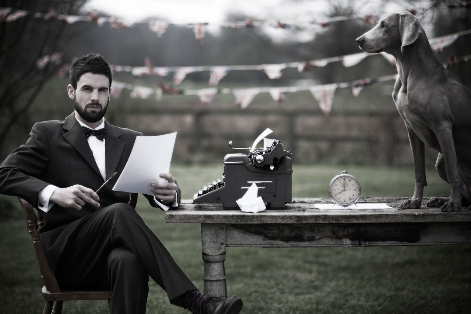 Portrait of a man sitting outside in a field at a table with a typewriter and a dog on the table. He is wearing a full suit and a bow tie. There is UK flag bunting hanging behind him. Shot by Norwich commercial, freelance portrait photographer Matt Keal.